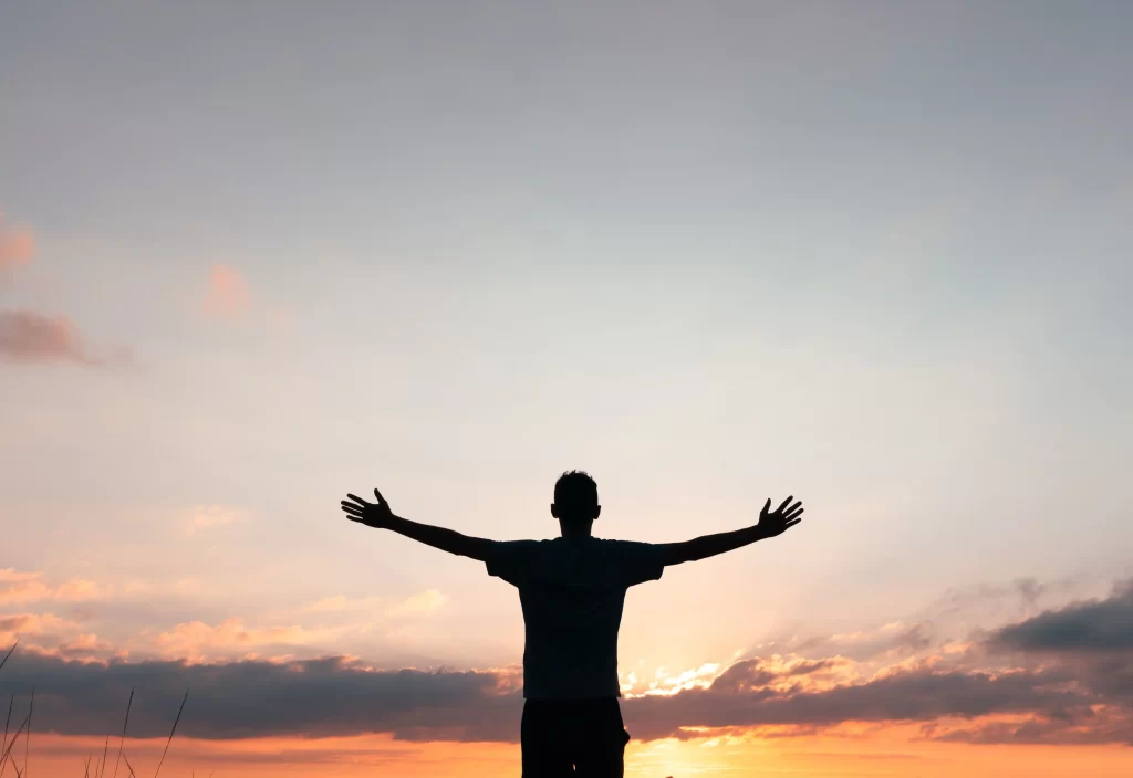 Muž s rozpřaženýma rukama a západem slunce.