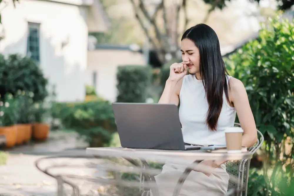 Žena sedí u notebooku na zahradě a drží si nos. 