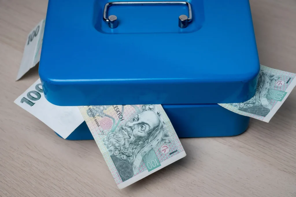 Mal&aacute; uzamykateln&aacute; modr&aacute; kovov&aacute; pokladnička, ze kter&eacute; trč&iacute; několik bankovek česk&eacute; měny v hodnotě 100 Kč. 