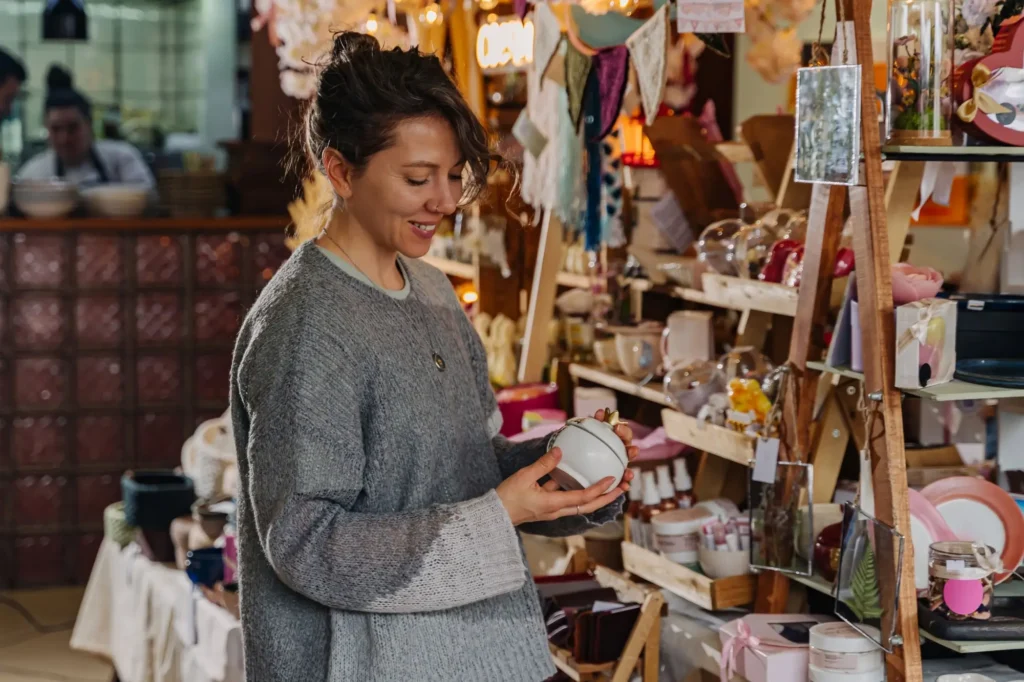 Žena v &scaron;ed&eacute;m svetru stoj&iacute; v obchodě s d&aacute;rkov&yacute;m zbož&iacute;m a prohl&iacute;ž&iacute; si malou keramickou d&oacute;zu