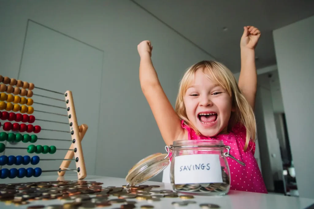 Holčička m&aacute; ruce v&iacute;tězoslavně nad hlavou, sed&iacute; u stolu s mincemi a sklenic&iacute;.