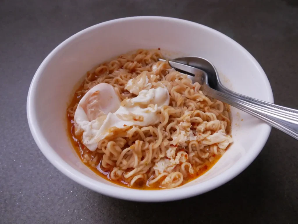 Miska instantn&iacute;ch nudl&iacute; s v&yacute;varem a vejcem, připraven&aacute; k j&iacute;dlu s vidličkou a lž&iacute;c&iacute; položenou na okraji.