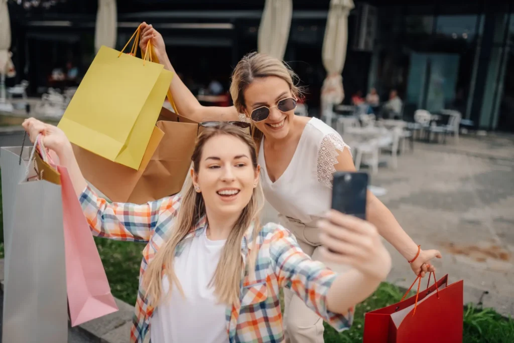 Dvě ženy venku p&oacute;zuj&iacute; se spoustou n&aacute;kupn&iacute;ch ta&scaron;ek a fot&iacute; si společn&eacute; selfie