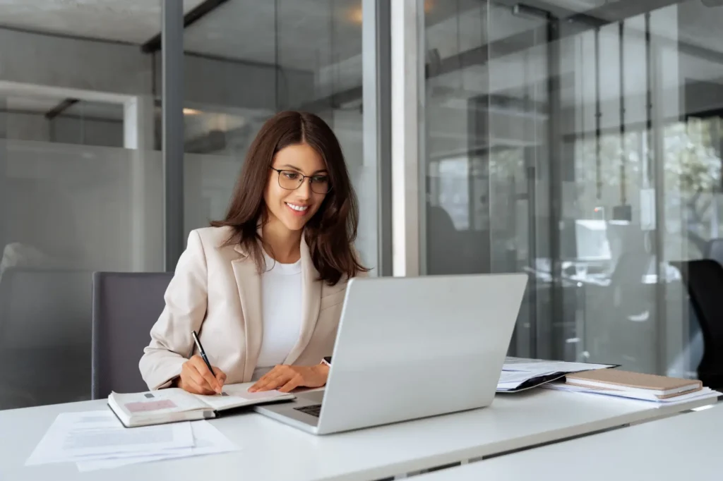 Usměvav&aacute; žena v br&yacute;l&iacute;ch sed&iacute; u stolu s notebookem, p&iacute;&scaron;e si pozn&aacute;mky a &uacute;častn&iacute; se online meetingu.