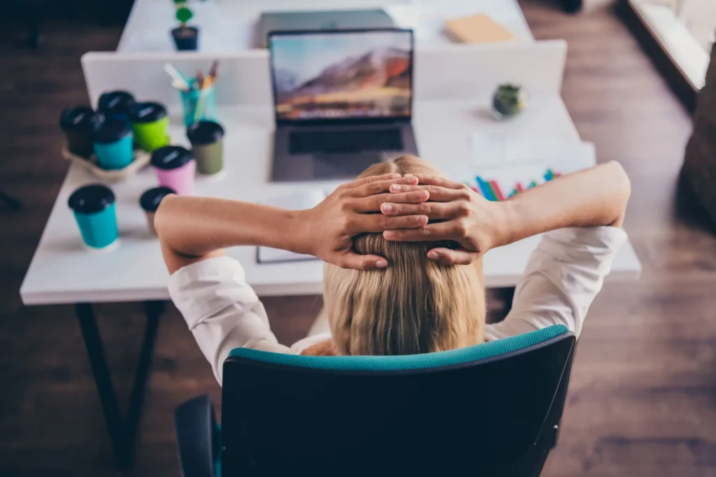 Žena sed&iacute; u pracovn&iacute;ho stolu, ruce m&aacute; založen&eacute; za hlavou a d&iacute;v&aacute; se na notebook.