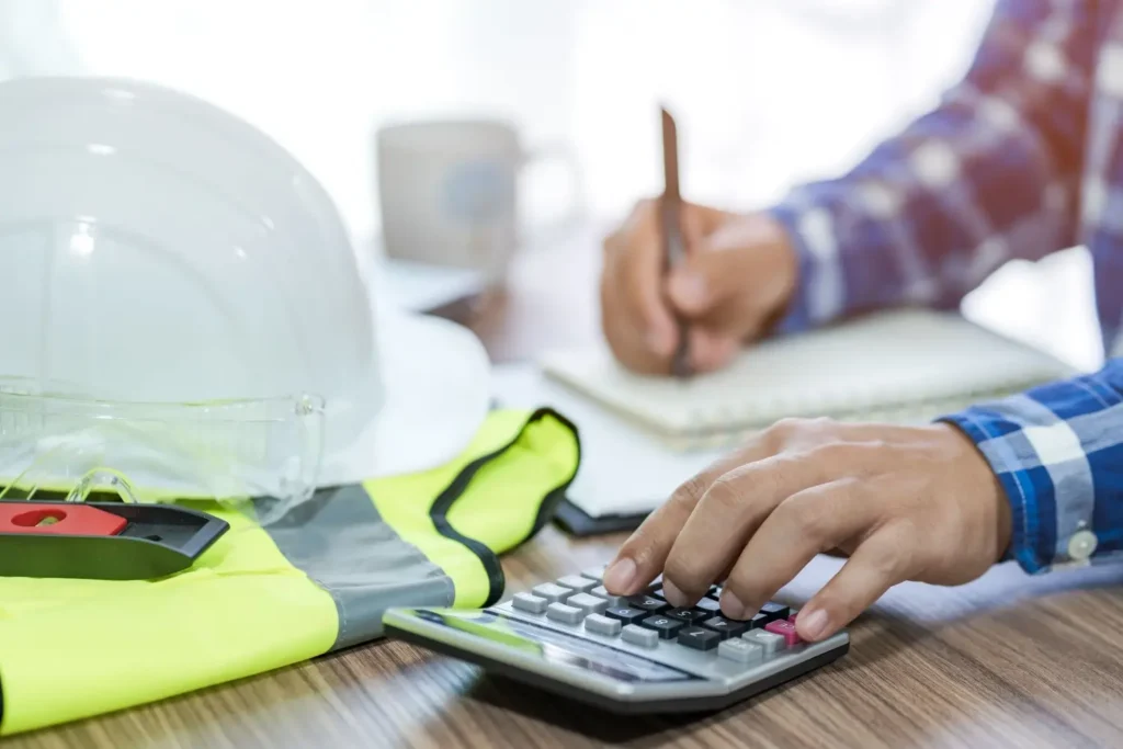 Zedn&iacute;k u stolu poč&iacute;t&aacute; n&aacute;klady stavby na kalkulačce, v popřed&iacute; lež&iacute; ochrann&aacute; přilba a reflexn&iacute; vesta