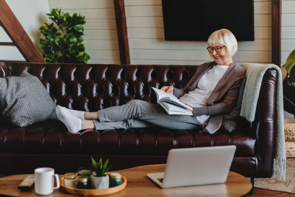 Usměvavá seniorka sedí pohodlně na pohovce s knihou a užívá si volný čas