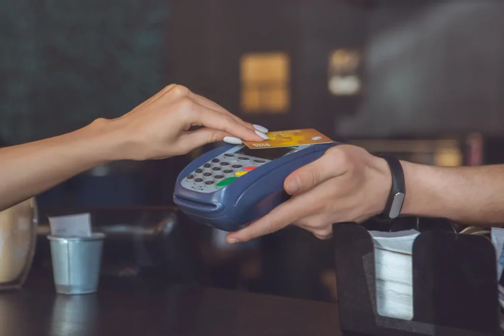 Detail ženské ruky přikládající zlatou platební kartu k terminálu drženému prodavačem