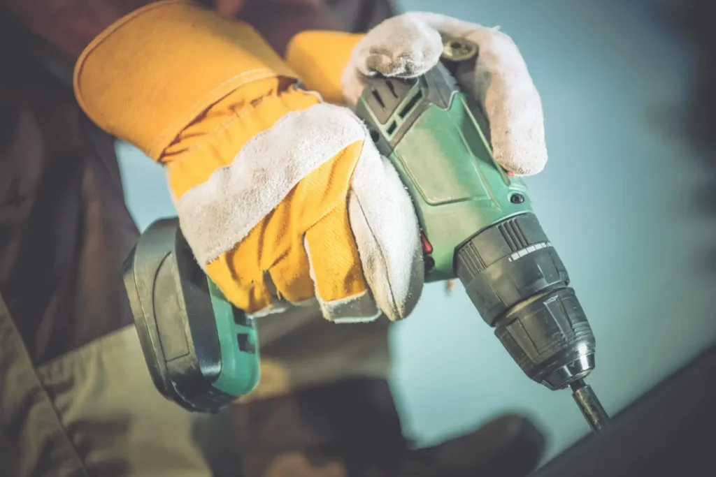 Detail rukou v pracovních rukavicích při práci s aku vrtačkou v dílně