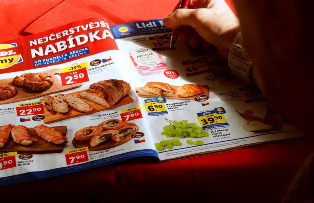 Osoba si prohl&iacute;ž&iacute; ti&scaron;těn&yacute; akčn&iacute; let&aacute;k česk&eacute;ho Lidlu s cenami pečiva, masa a ovoce