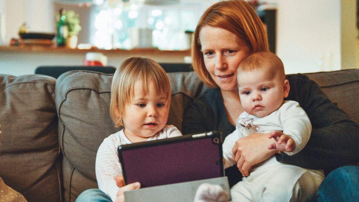 two babies and woman sitting on sofa while holding baby and watching on tablet