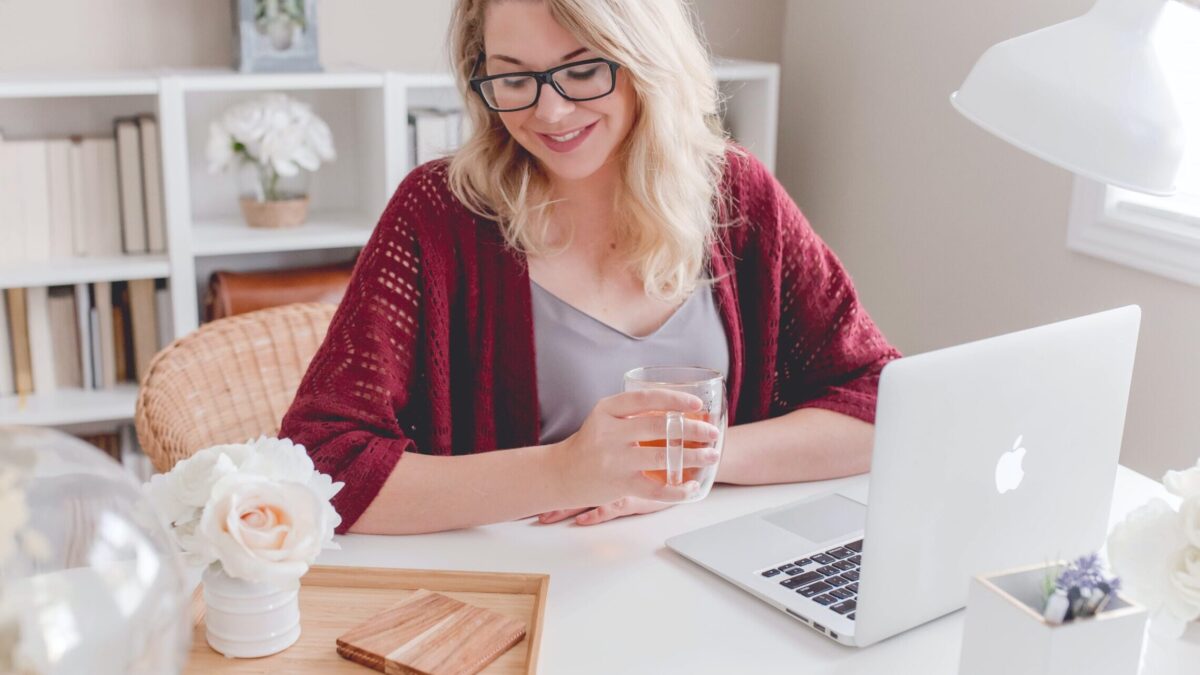 woman smiling holding glass mug sitting beside table with MacBook