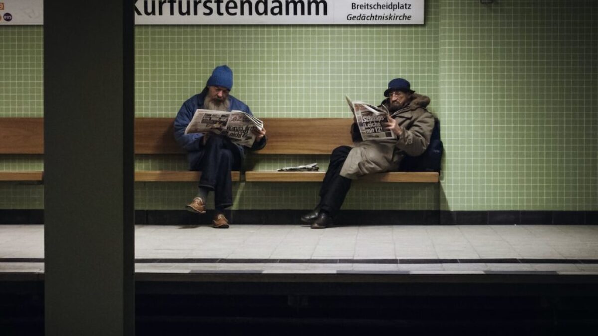 two men sitting on brown wooden bench reading magazines