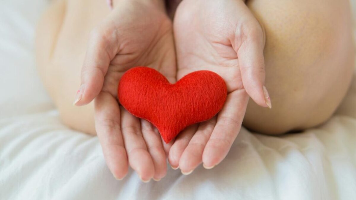 Unrecognizable female sitting with bare legs on white sheet with small red heart in hands in light room in daytime