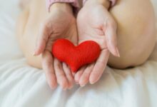 Unrecognizable female sitting with bare legs on white sheet with small red heart in hands in light room in daytime