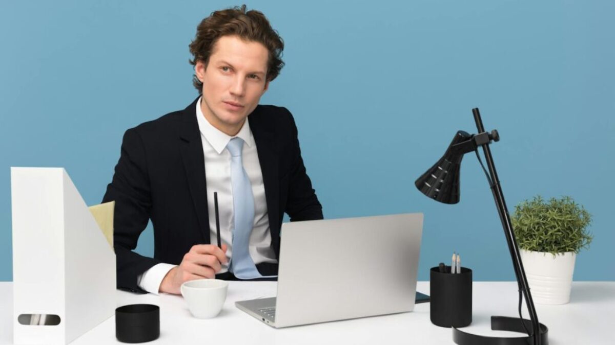 A focused businessman in a suit works at his tidy desk with a laptop, plant, and lamp.