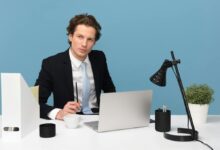 A focused businessman in a suit works at his tidy desk with a laptop, plant, and lamp.