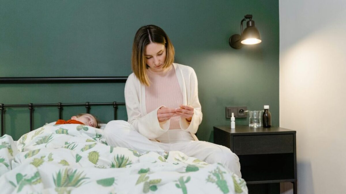 A mother beside her sick child holding medicine in a cozy bedroom setting.