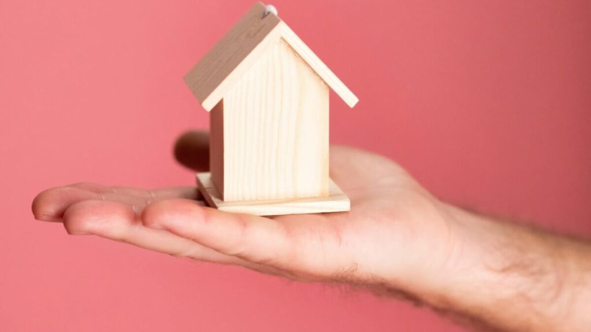 Wooden house model in hand symbolizing real estate concepts on pink background.