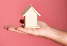 Wooden house model in hand symbolizing real estate concepts on pink background.