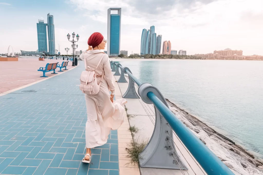 Shutterstock_2287888959 - ByznysDeník Žena v hidžábu a dlouhém oblečení kráčí podél promenády s výhledem na moderní mrakodrapy.