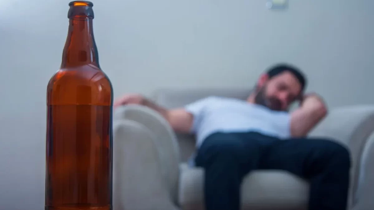 Muž sedí na gauči, v popředí láhev alkoholu.
