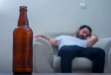Muž sedí na gauči, v popředí láhev alkoholu.
