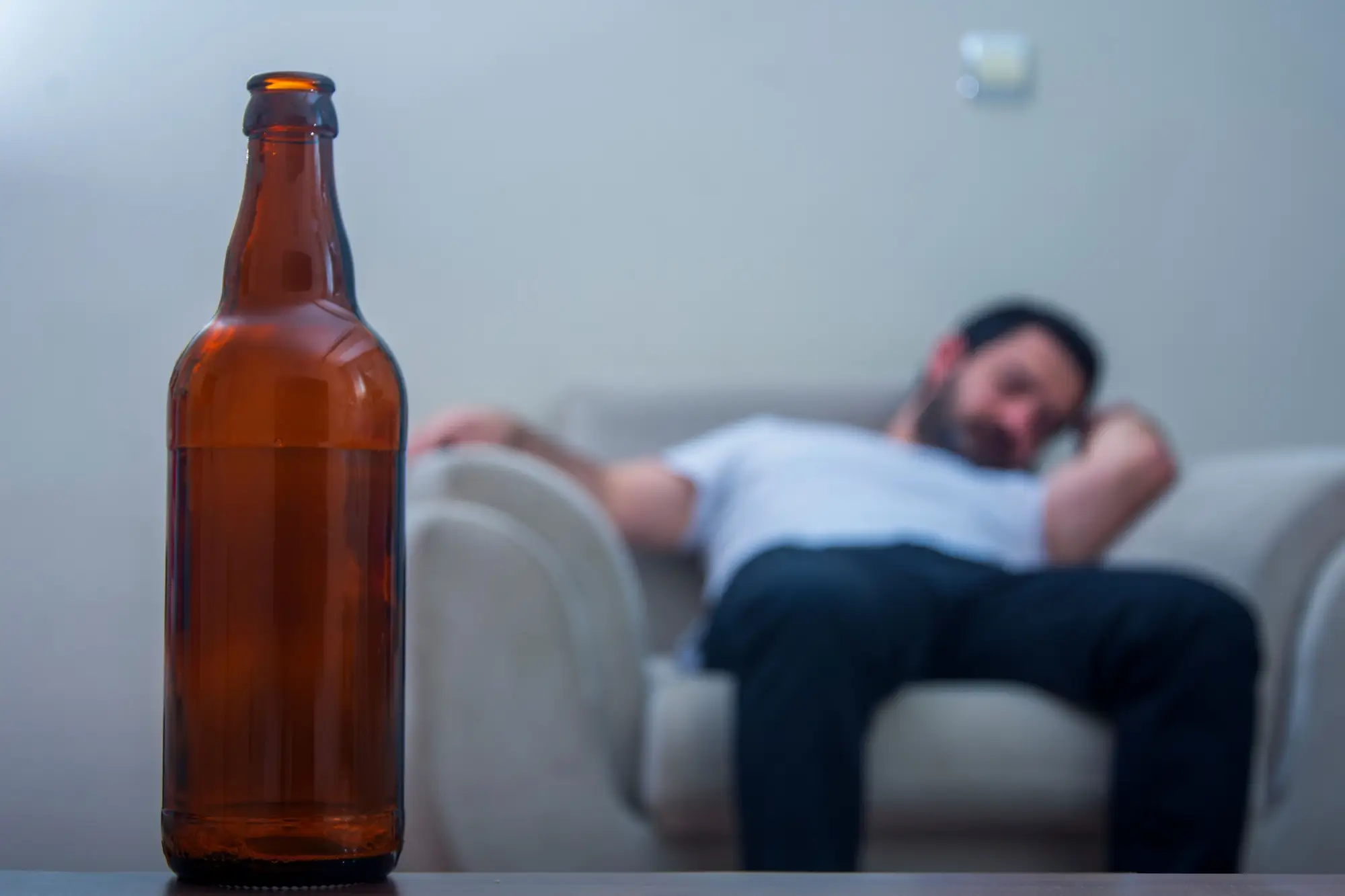Muž sedí na gauči, v popředí láhev alkoholu.