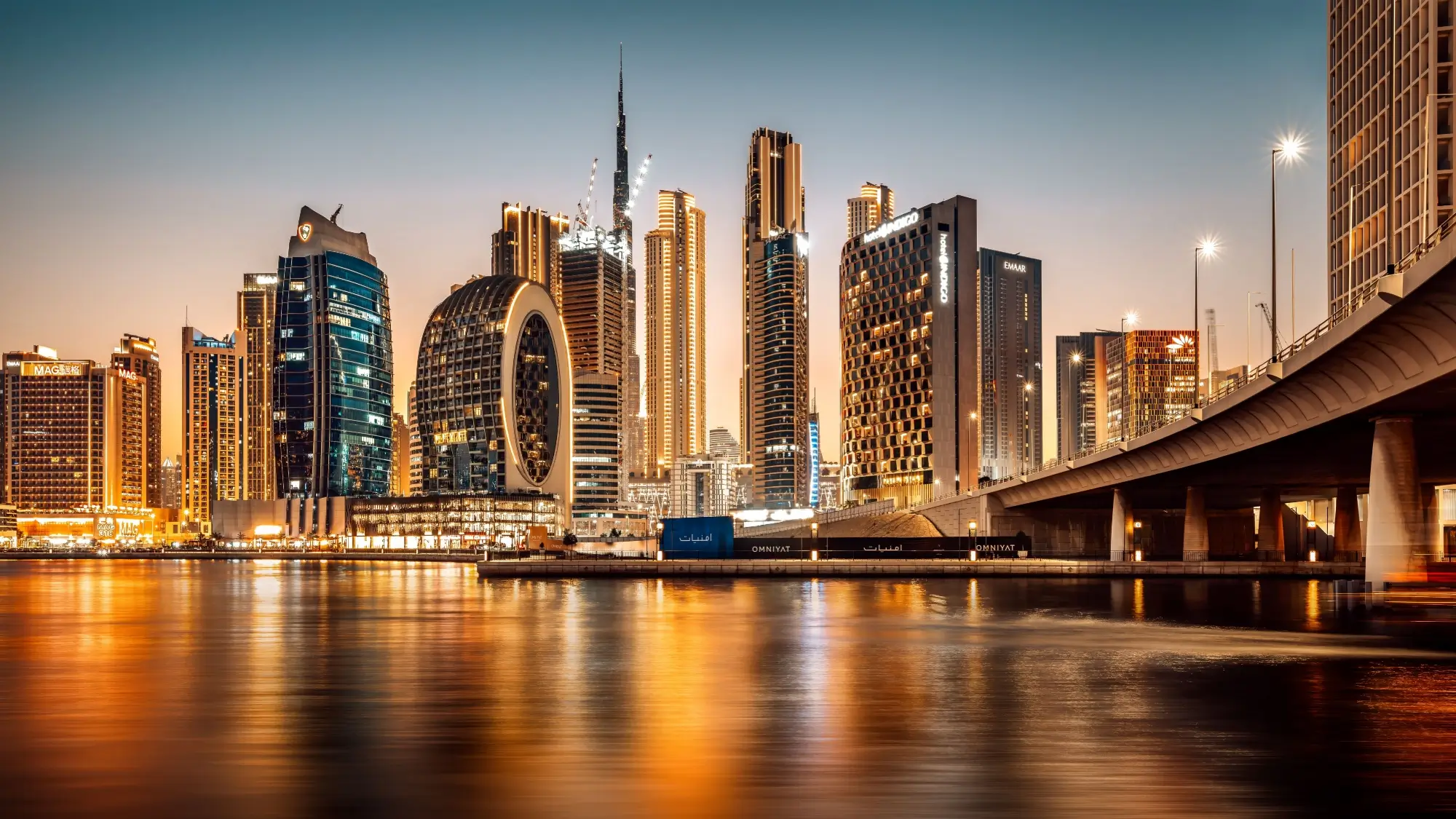 Noční pohled na futuristické panorama Dubaje s rozsvícenými mrakodrapy a odrazem ve vodní hladině.