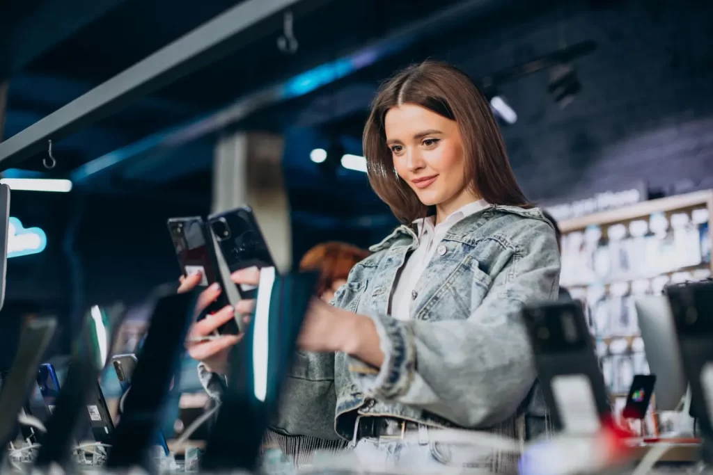 Za čas možná budete pořizovat novější telefon. - ByznysDeník Žena v obchodě vybírá nový mobil.