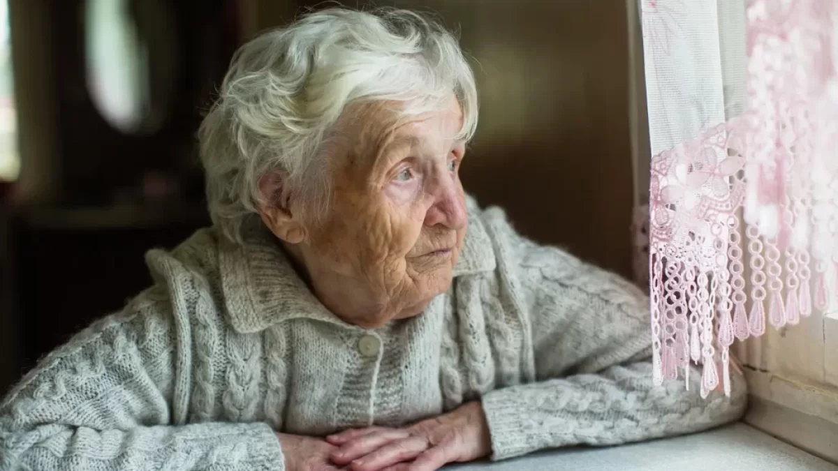 Seniorka sedí u stolu a dívá se z okna.