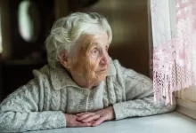 Seniorka sedí u stolu a dívá se z okna.