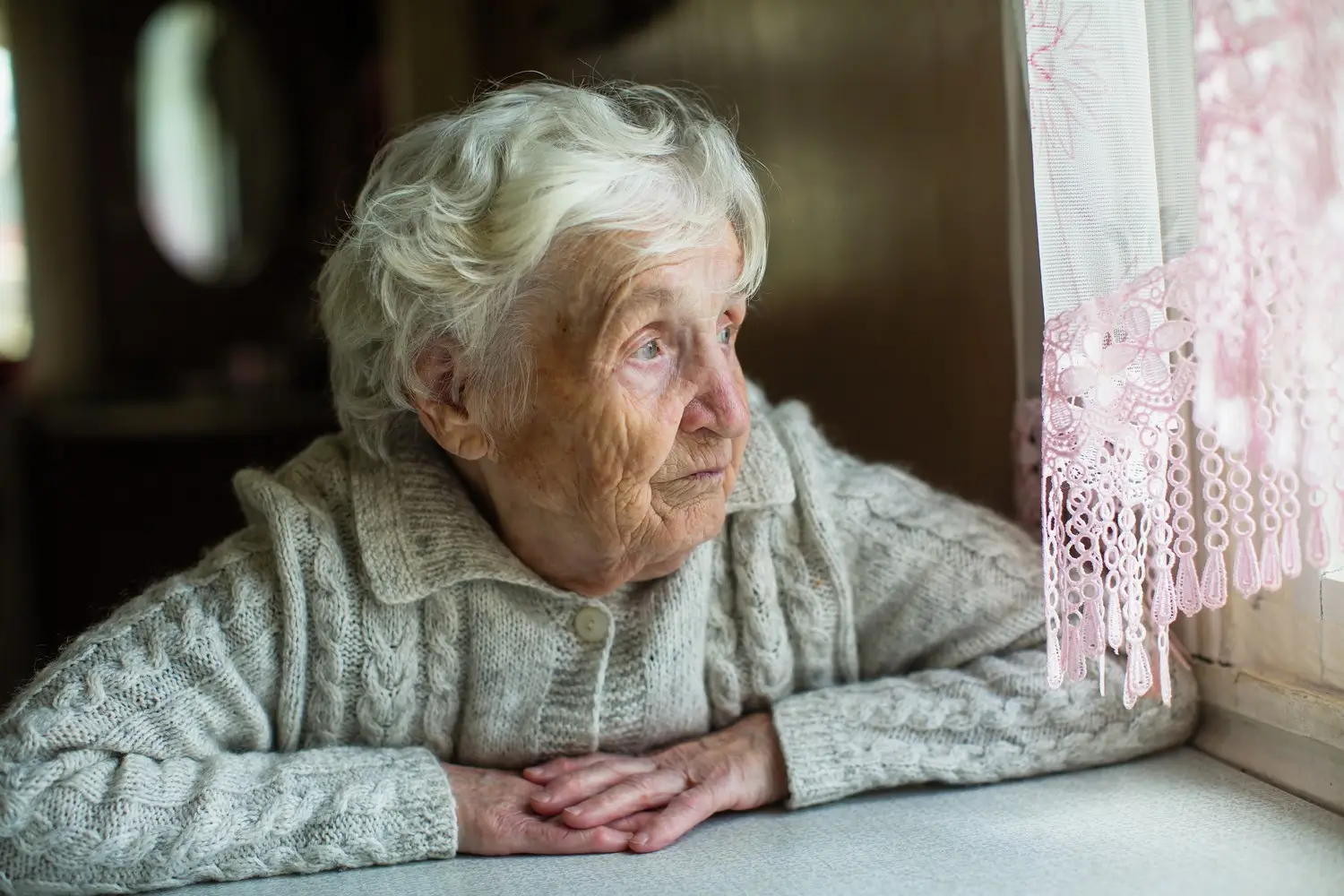 Seniorka sedí u stolu a dívá se z okna.