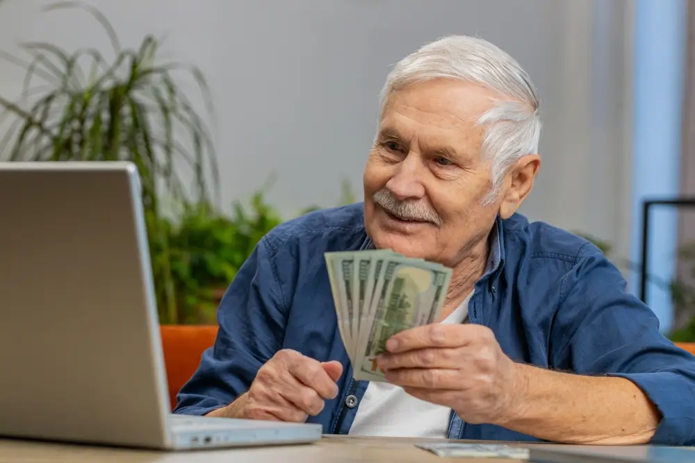 shutterstock_2524684269 - ByznysDeník Starší muž se usmívá na notebook a drží v ruce bankovky.