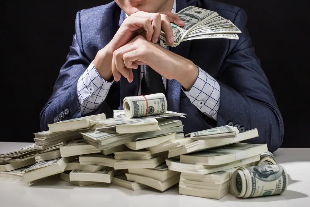 shutterstock_710867164 - ByznysDeník Muž sedí u stolu, na němž je plno bankovek.