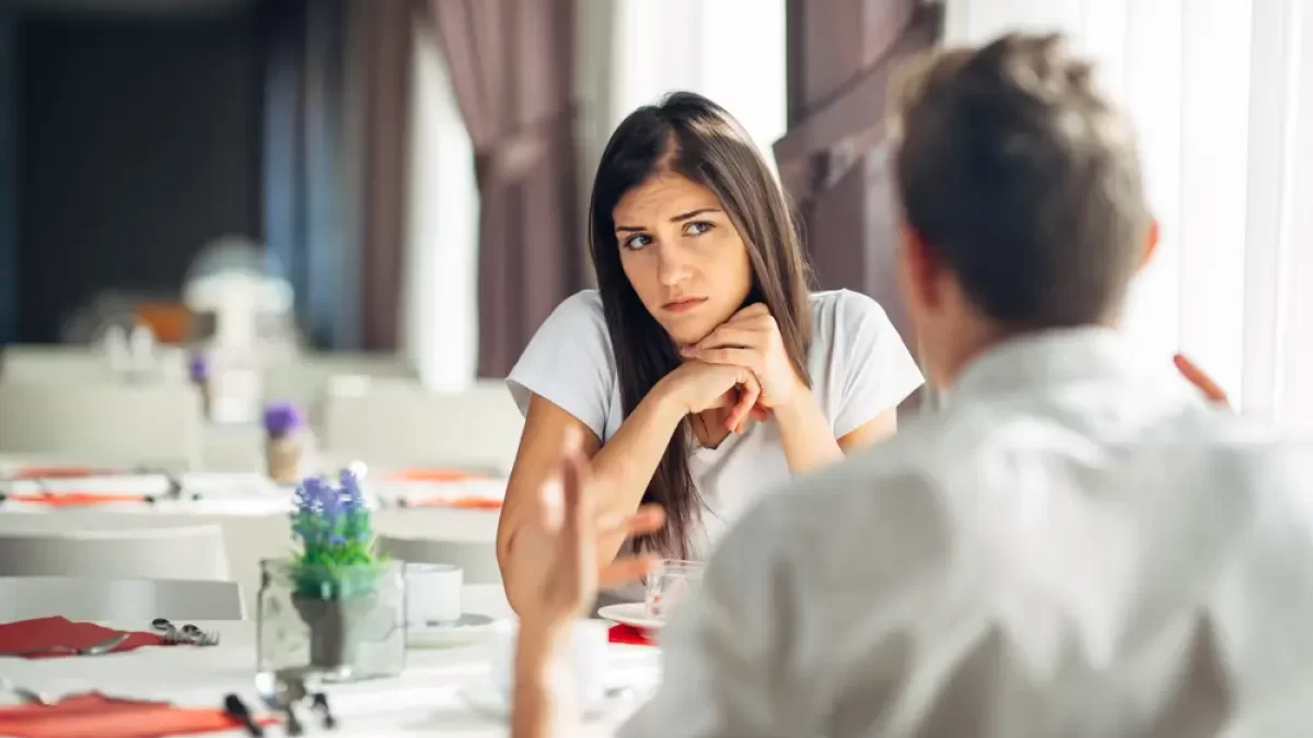 Žena sedí naproti muži v restauraci a podezřívavě se na něj dívá.