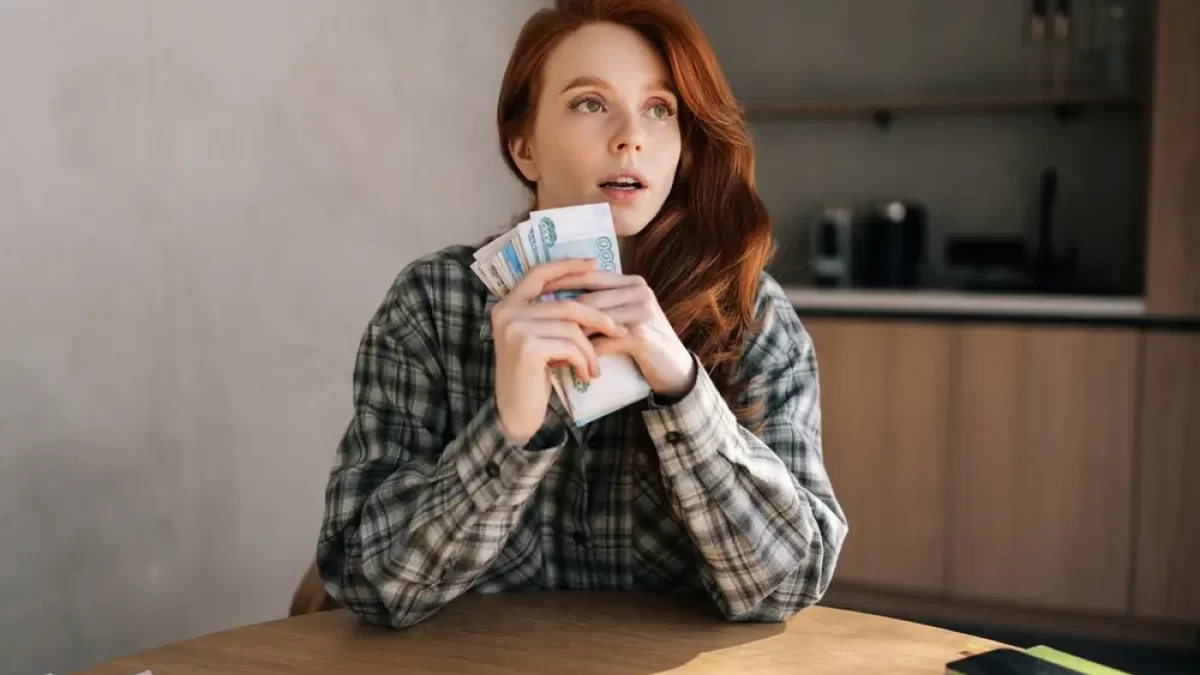 Žena sedí u stolu, v ruce drží balík bankovek.