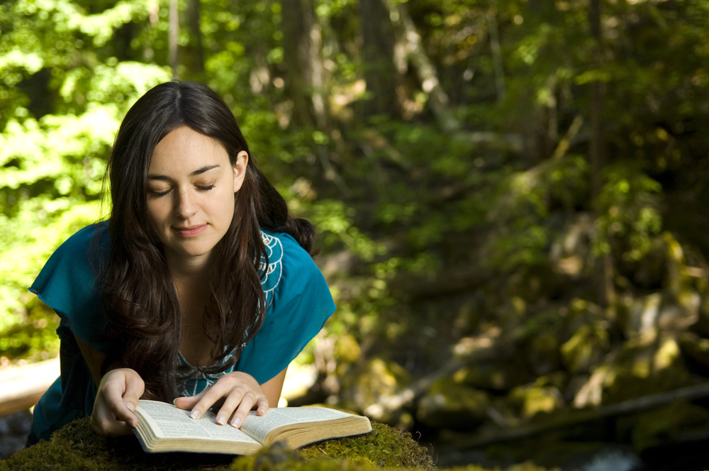 YoungWomanReadingBibleByStreamInSummer - ByznysDeník Žena si čte knihu uprostřed lesa.