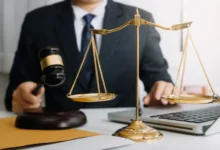 Muž v obleku drží kladívko u stolu vedle zlatých vah a otevřeného notebooku