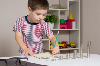 Malý chlapec si zapisuje poznámky do sešitu a zároveň před sebou počítá mince uspořádané do sloupců podle výšky.
