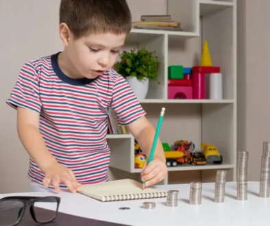 Malý chlapec si zapisuje poznámky do sešitu a zároveň před sebou počítá mince uspořádané do sloupců podle výšky.