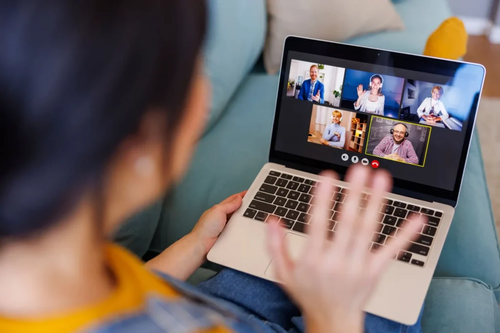 - ByznysDeník Žena sedí na gauči s notebookem a účastní se videokonference s pěti lidmi, přátelsky jim mává