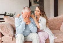 Smutný starší muž sedí na gauči a mladší žena jej utišuje