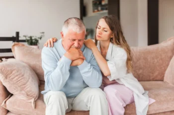 Smutný starší muž sedí na gauči a mladší žena jej utišuje