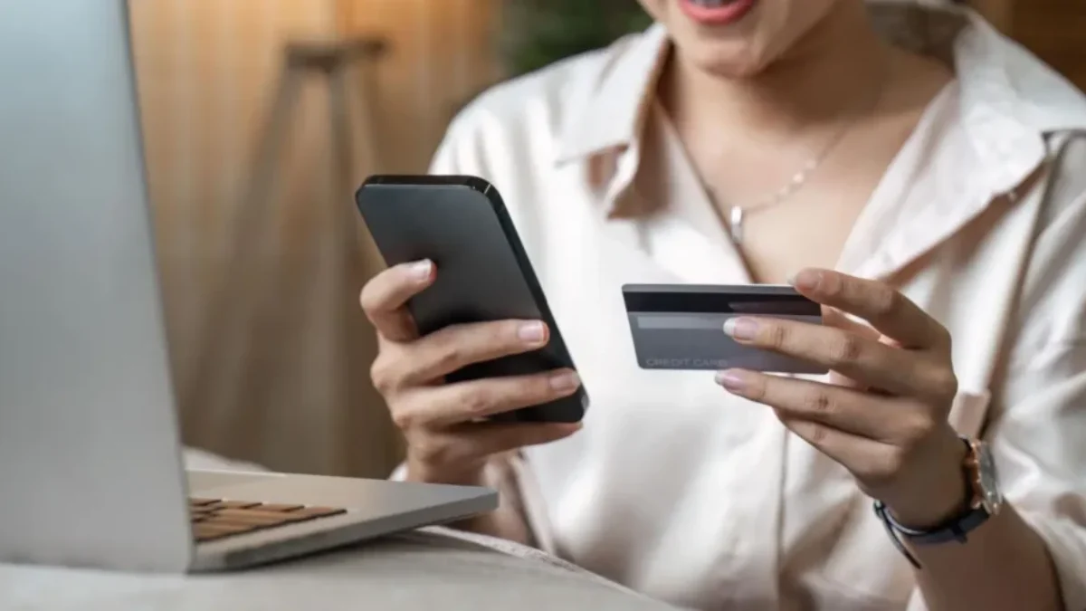 Žena drží v ruce mobilní telefon a platební kartu před otevřeným notebookem