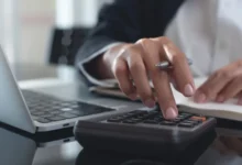 Detail ruky počítající výdaje na kalkulačce u notebooku, ilustrační foto domácího rozpočtu