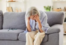 Seniorka sedí na pohovce a drží se za hlavu.