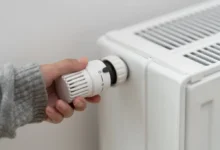 Detail ruky, která otáčí hlavicí termostatu na bílém radiátoru, za účelem snížení teploty v místnosti.