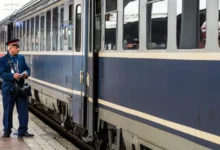 Průvodčí ve služební uniformě, který stojí na nástupišti vedle modrého osobního vlaku.