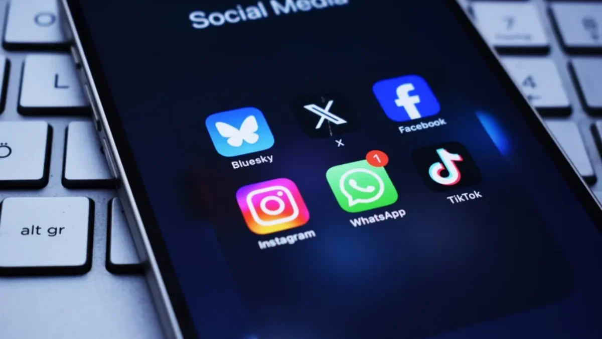 Na obrazovce telefonu je otevřená složka „Social Media“ se šesti ikonami aplikací: Bluesky, X (dříve Twitter), Facebook, Instagram, WhatsApp.